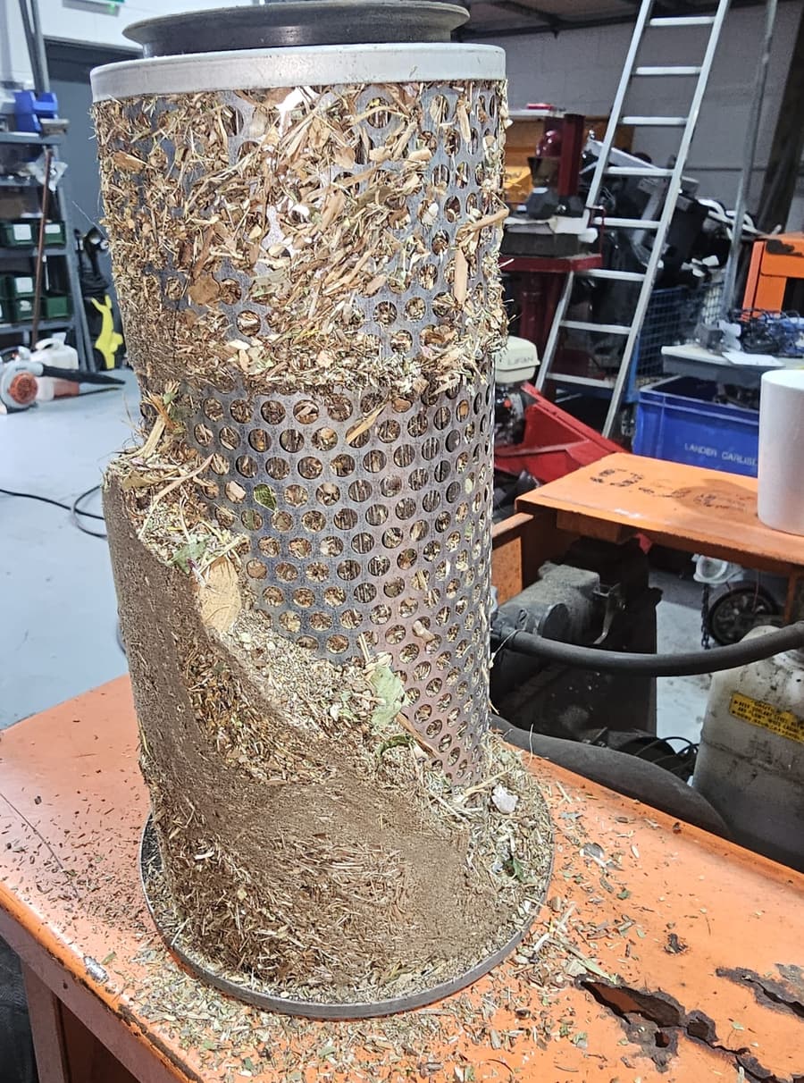 An air filter element on a workbench, its perforated steel body packed solid with dried grass, chaff and leaf dust
