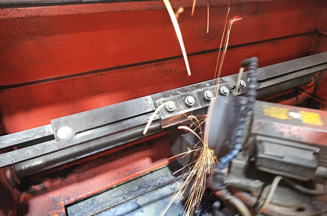 A chipper blade clamped along the bed of the Chippermaster knife grinder, the wheel part-way down its edge, sparks coming off